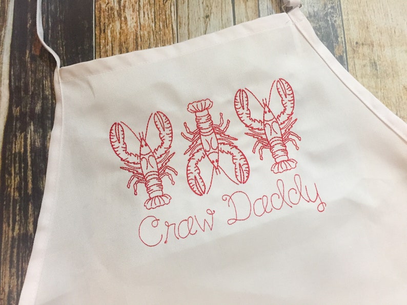 May include: White apron with red embroidered lobsters and the text "Crew Daddy".