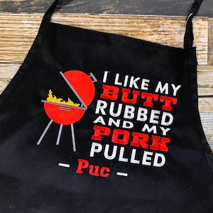 May include: Black apron with the text "I LIKE MY BUTT RUBBED AND MY PORK PULLED - Puc" in white and red lettering. A red grill with flames is embroidered on the left side. The apron has a neck loop and waist ties.