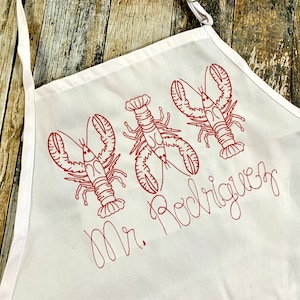 May include: White apron with red embroidered lobsters and the text "Mr. Rodriguez".