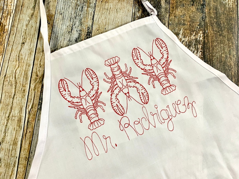 May include: White apron with red embroidered lobsters and the text "Mr. Rodriguez".