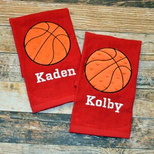 May include: Two red towels with embroidered basketballs and names. The towels feature a white basketball with orange stitching and a white embroidered name. The names on the towels are "Kaden" and "Kolby".