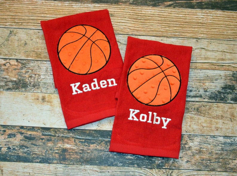 May include: Two red towels with embroidered basketballs and names. The towels feature a white basketball with orange stitching and a white embroidered name. The names on the towels are "Kaden" and "Kolby".