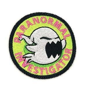 May include: A round, green and black embroidered patch with a white ghost with a black mouth and the words "Paranormal Investigator" in pink.