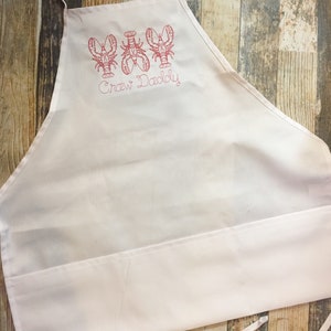 May include: White kitchen apron with a red embroidered design of three lobsters and the text "Craw Daddy".