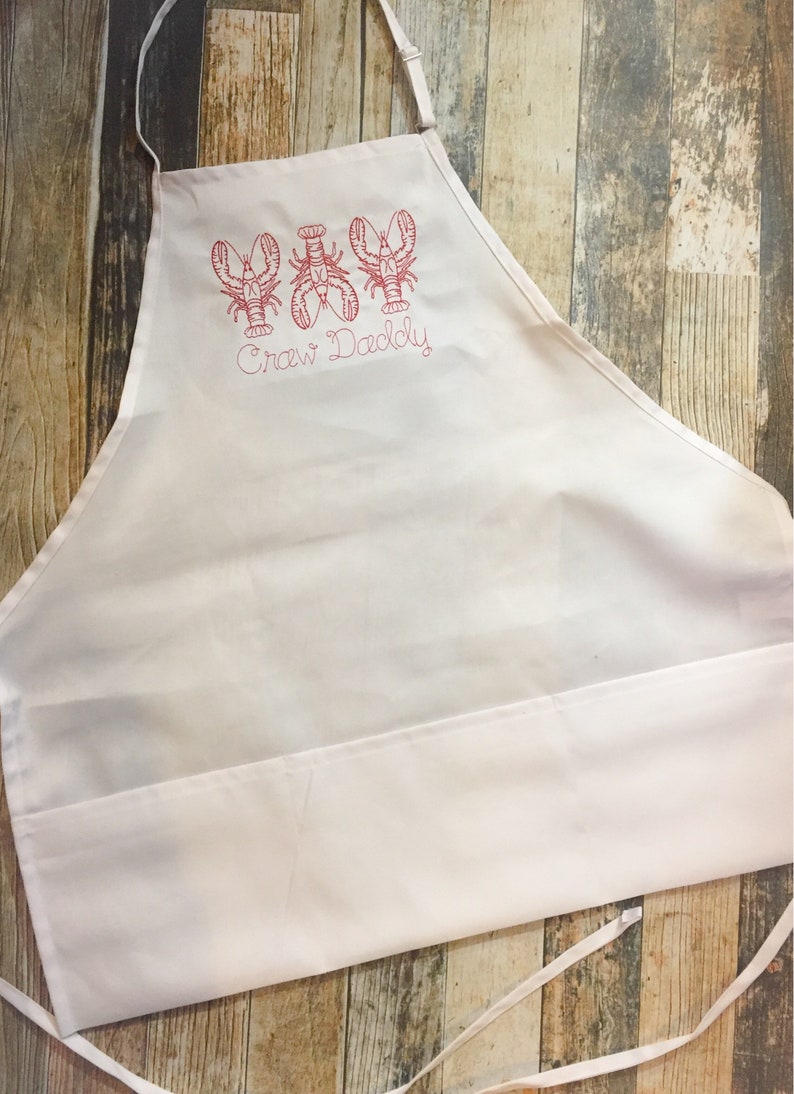May include: White kitchen apron with a red embroidered design of three lobsters and the text "Craw Daddy".