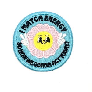 May include: A round embroidered patch with a light blue border and the text "I MATCH ENERGY SO HOW WE GONNA ACT TODAY!". The patch features a pink flower with a yellow center and two white peace signs.