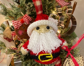 May include: A crocheted Santa Claus ornament with a red hat, white beard, and a black belt with a yellow buckle. The ornament is hanging on a Christmas tree with other decorations.