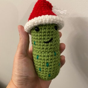 May include: A handmade, crocheted pickle with a smiling face, black eyes, and teal accents. The pickle wears a red and white Santa hat, suggesting a holiday theme. The item is held in a hand.