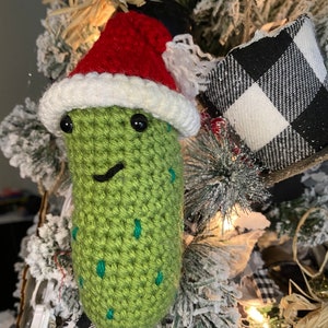 May include: A handmade, green crocheted pickle Christmas ornament wearing a red Santa hat with white trim. The pickle has black eyes and a smiling mouth. The ornament is decorated with green spots and is hanging on a Christmas tree.