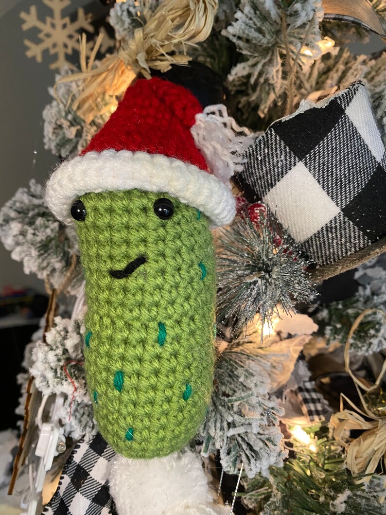 May include: A handmade, green crocheted pickle Christmas ornament wearing a red Santa hat with white trim. The pickle has black eyes and a smiling mouth. The ornament is decorated with green spots and is hanging on a Christmas tree.