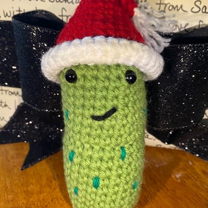 May include: A crocheted green pickle wearing a red and white Santa hat. The pickle has black eyes and a stitched smile, with teal accents. The text "Pattern only" is at the bottom.