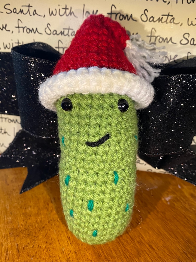 May include: A handmade, crocheted pickle wearing a red and white Santa hat. The pickle is green with small teal accents and black eyes, and a smiling mouth. The background includes a black bow and handwritten text.