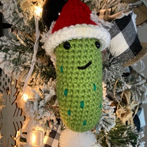 May include: A handmade, green crocheted pickle Christmas ornament wearing a red and white Santa hat. The pickle has black eyes and a smiling mouth. The ornament is decorated with small teal stitches and a white pom-pom.