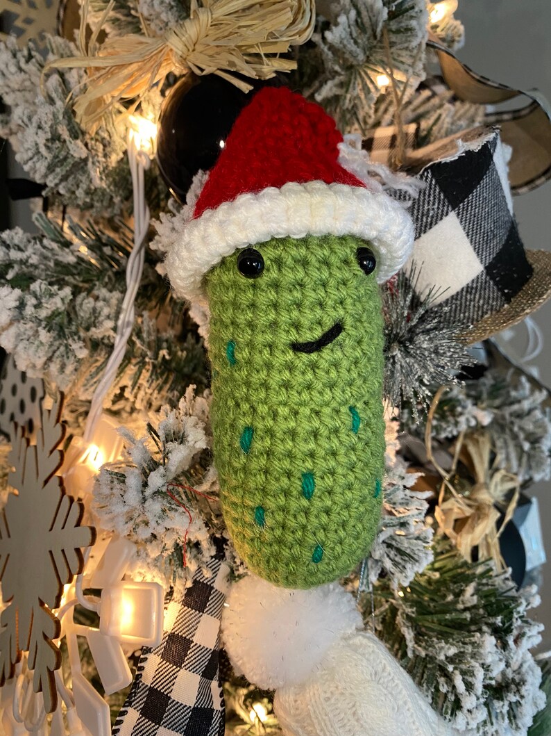 May include: A handmade, green crocheted pickle Christmas ornament wearing a red and white Santa hat. The pickle has black eyes and a smiling mouth. The ornament is decorated with small teal stitches and a white pom-pom.