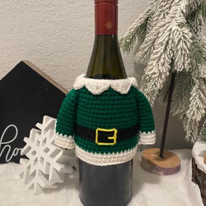 May include: A green and white crocheted wine bottle cover, designed to look like an elf's attire. It features a white collar, a black belt with a gold buckle, and white cuffs. The wine bottle has a red top with a gold star design.
