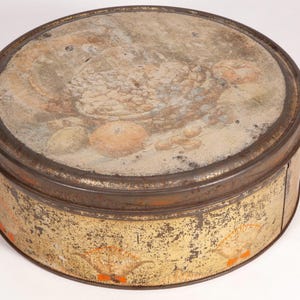 May include: A round, vintage metal tin with a lid featuring a faded still-life painting of fruit. The tin has a gold and brown color scheme with signs of wear and age, adding to its antique charm. The lid's artwork includes oranges and other fruits.