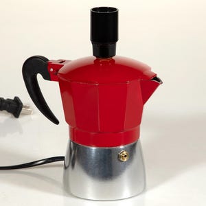 May include: A red and silver electric stovetop espresso maker with a black handle and a black power cord. The coffee maker has a black top and a silver base. The power cord is plugged into an electrical outlet.