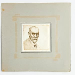 May include: A sepia-toned portrait print of a man with a bald head and a beard, wearing a collared shirt and tie. The artwork is mounted on a light gray square mat board with visible tape marks at the corners and a signature at the bottom edge.