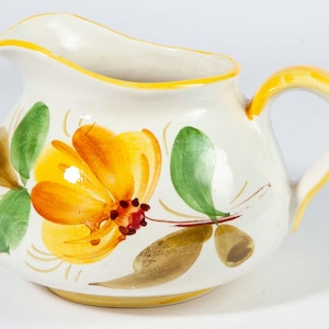 Op de afbeelding: Een witte keramische kan met een gele handgreep en rand. De kan is versierd met een grote geel-oranje bloem, groene bladeren en bruine accenten. Een decoratieve bloemenkan.