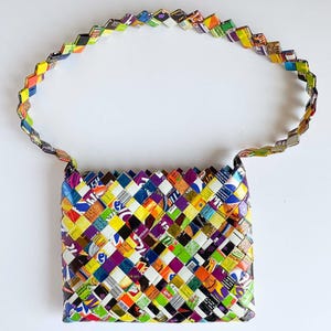 May include: A rectangular handbag constructed from woven strips of colorful, printed candy wrappers. The bag features a single, matching woven strap and a multi-colored geometric pattern throughout, showcasing various shades of green, purple, yellow, and blue.