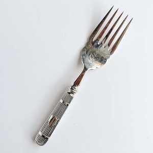 May include: A silver-toned metal fork featuring a four-tined head with an ornate, engraved floral pattern at the base. The handle is cylindrical with vertical ribbed detailing and a small, smooth shield-shaped emblem near the bottom end.