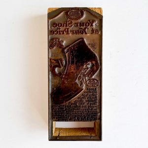 May include: A vintage shoe printmaking block with the text "In Your Shoe's Juice" and an illustration of a shoe. The block is made of metal and wood, with the shoe illustration in the center.