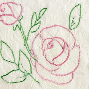 May include: A piece of off-white, textured fabric featuring hand-stitched embroidery of two roses. The rose petals are outlined in pink thread, while the stems and leaves are detailed with green thread, creating a floral pattern on the cloth.