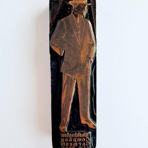 May include: A rectangular metal printing block featuring an engraved illustration of a person wearing a suit and hat. The base of the block displays the embossed text Washington Company Garments New York in a mirrored, inverted orientation.