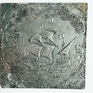 May include: A square, silver-colored metal relief depicting a sailing ship on water, surrounded by a decorative border. The ship has multiple sails and flags, with a detailed depiction of the sea and sky.