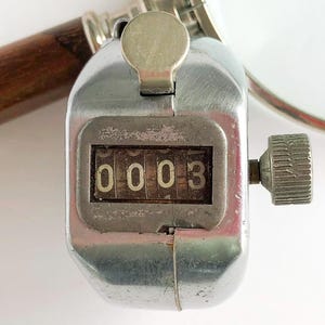 May include: A handheld silver-toned metal tally counter featuring a mechanical display showing the number 0003. The device includes a top push-button actuator and a side-mounted knurled reset knob, with a wooden handle visible in the background.