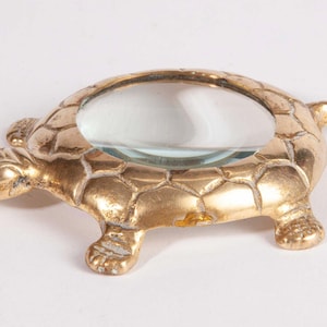 May include: A gold-toned metal turtle-shaped magnifying glass featuring a polished finish and a textured shell pattern. The circular glass lens is set into the center of the turtle's back, with a head, four legs, and a tail extending outward.