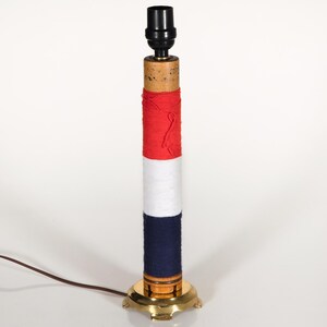 May include: A cylindrical table lamp base featuring a wooden core wrapped in horizontal bands of red, white, and dark blue thread. The lamp stands on a circular brass base and is topped with a black socket, accompanied by a brown electrical cord.