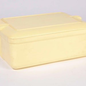 Może przedstawiać: Prostokątne, jasnożółte pudełko do przechowywania z pasującą pokrywą. Pudełko ma zaokrąglone rogi i lekko teksturowaną powierzchnię. Pokrywa ma wpuszczony panel i małe, podniesione detale po bokach. Pudełko ma około 30 cm długości.