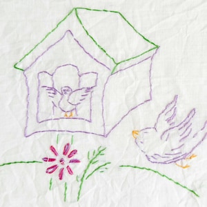 May include: A white fabric panel featuring purple and green embroidery of a birdhouse with a bird inside, a flying bird to the right, and a small pink flower at the base. The design uses simple outline stitching on a textured white background.
