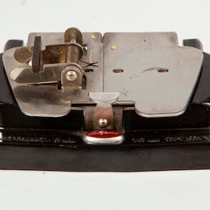 May include: A vintage black film splicer featuring a glossy base and a metallic top plate with a hinged clamp mechanism. The device includes a small red oval emblem with the word Hollywood printed in white script on the front center section.