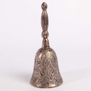May include: A metallic hand bell with a polished bronze finish. The bell features an intricate embossed pattern of holly leaves and berries covering the exterior surface. It has a slender, vertical handle with a rounded, decorative finial.