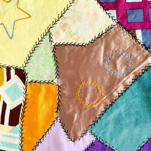 May include: A colorful patchwork textile with various fabric squares stitched together with black thread. The design includes yellow, orange, brown, purple, and teal fabrics, with embroidered star and circle details.
