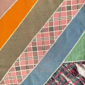 Op de afbeelding: Een patchwork textiel met diagonale strepen in verschillende kleuren, waaronder oranje, bruin, roze ruit, blauw en groen. Witte stiksels scheiden de strepen. De rechter benedenhoek heeft een gefranjerde ruitsectie.