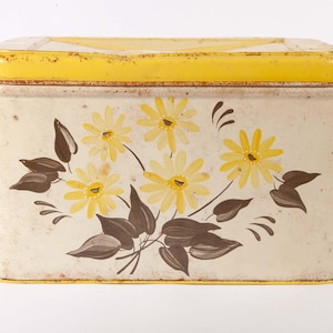 May include: A rectangular metal storage tin with a cream-colored body and a bright yellow lid. The front features a painted floral design with yellow daisy-like flowers and dark brown leaves. The edges of the lid and base show signs of rust.