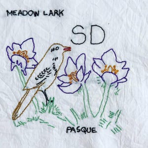 Pode incluir: Tecido branco bordado com um cotovia, flores roxas e brancas e o texto "MEADOW LARK", "SD" e "PASQUE". O pássaro é castanho, branco e preto, com um bico vermelho. As flores têm contornos roxos e centros amarelos.