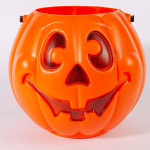 May include: A bright orange plastic pumpkin bucket featuring a molded jack-o'-lantern face with cut-out eyes, a triangular nose, and a wide, toothy grin. The container has a rounded, ribbed body and small black attachment points for a handle.