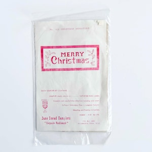 Może przedstawiać: Zestaw do haftu bożonarodzeniowego w przezroczystej plastikowej torbie. Opakowanie jest w kolorze złamanej bieli z czerwonym banerem z napisem "MERRY Christmas". Zestaw pochodzi od Jane Snead Samplers, ze słowami "Keepsake Handiwork".