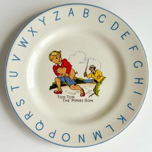 Puede incluir: Un plato blanco vintage con un borde azul y letras del alfabeto azules alrededor del borde. El plato presenta una ilustración colorida de un niño que lleva un cerdo, con un hombre con una chaqueta amarilla. El texto dice "Tom Tom The Pipers Son."