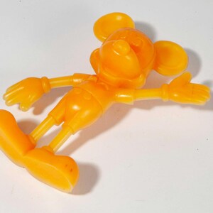 May include: An orange plastic figurine of a cartoon mouse with large ears, gloves, and shoes. The mouse is lying on its side with its arms outstretched. The figurine is a solid color and appears to be a toy or collectible item.