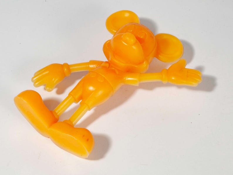 May include: An orange plastic figurine of a cartoon mouse with large ears, gloves, and shoes. The mouse is lying on its side with its arms outstretched. The figurine is a solid color and appears to be a toy or collectible item.