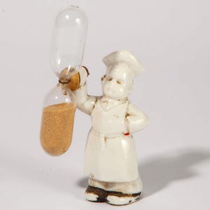 May include: A white ceramic figurine of a chef holding a clear glass hourglass filled with tan sand. The chef is wearing a tall hat, apron, and has a stern expression. The hourglass is approximately 4 inches tall.