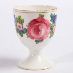 May include: A white porcelain egg cup featuring a hand-painted floral pattern with a large central pink rose, smaller purple and blue flowers, and green leaves. The rim of the cup is accented with a thin gold line, and it stands on a pedestal base.