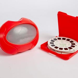 May include: A red View-Master stereoscope viewer with a blue advance lever and a translucent white lens cover, accompanied by a red plastic storage case containing a stack of circular white film reels with multiple image frames.