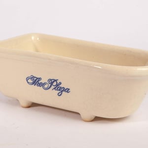 May include: A cream-coloured ceramic dish shaped like a miniature clawfoot bathtub. The exterior features the text "The Plaza" printed in a blue script font. The vessel has four small feet and a smooth, glossy finish on the interior and exterior.