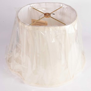 Puede incluir: Una pantalla de lámpara color crema con un diseño plisado, envuelta en plástico transparente. La pantalla tiene un marco de metal dorado y una etiqueta adjunta. La forma de la pantalla es clásica, adecuada para varias bases de lámparas.